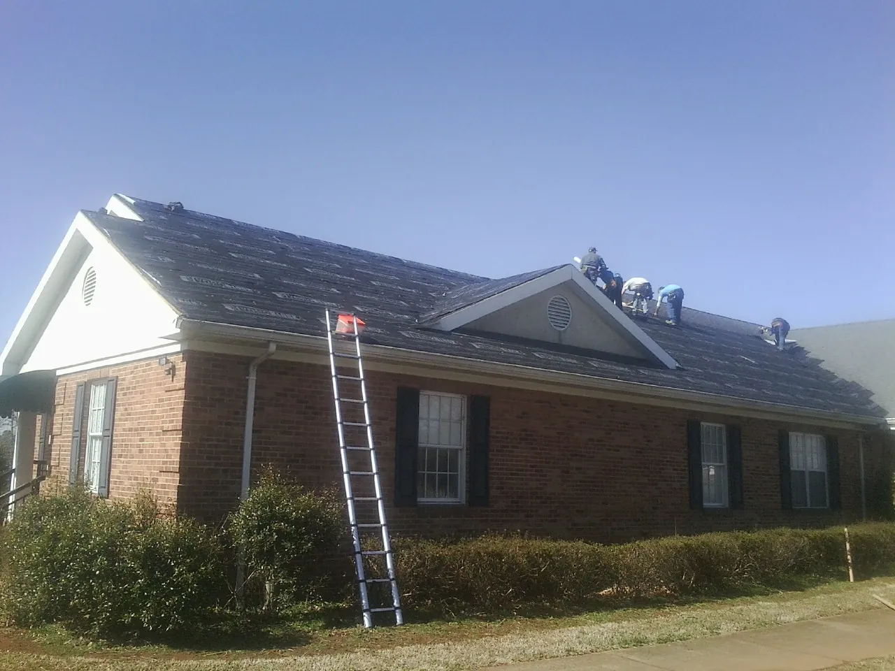 Crew tearing shingles off a brick ranch roof with a ladder set — Emergency Roof Repair work by our crew in Le Sueur