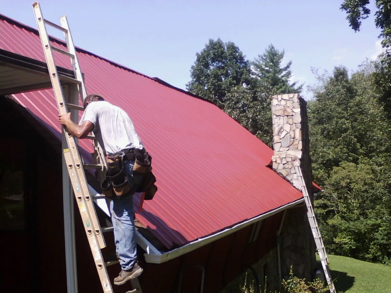 Roofer on a ladder beside a red metal roof and stone chimney — Soffit & Fascia Repair by Roofing Brothers in Le Sueur